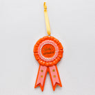 A novelty ornament shaped like a vintage award ribbon rosette, featuring a bright orange outer rosette, a pink inner rosette, white faux pearls around the center, and two pink ribbon tails with gold stars. The center reads 'World's Greatest' in gold script.