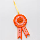 An orange and pink rosette-shaped ornament with a gold hanging ribbon, featuring the words 'World's Greatest' on the center label.
