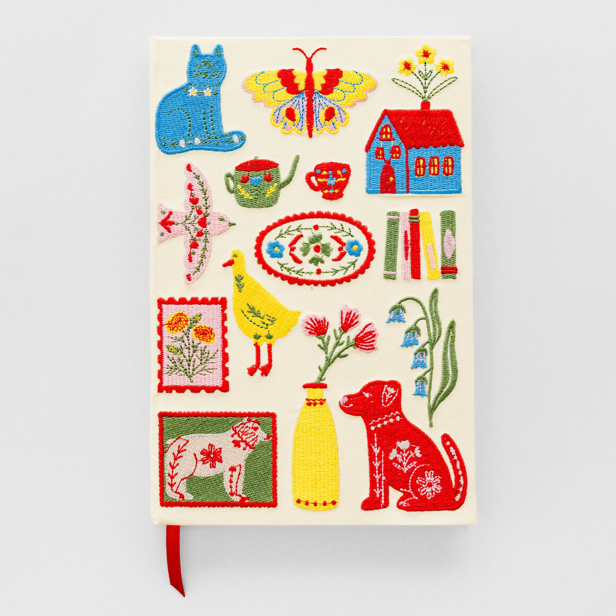 A hardcover journal with a cream-colored cover decorated with colorful embroidered folk-art style illustrations, including a blue cat, butterfly, house, flowers, teacups, books, and a red dog. A red ribbon bookmark is visible at the bottom left, set against a light gray background.