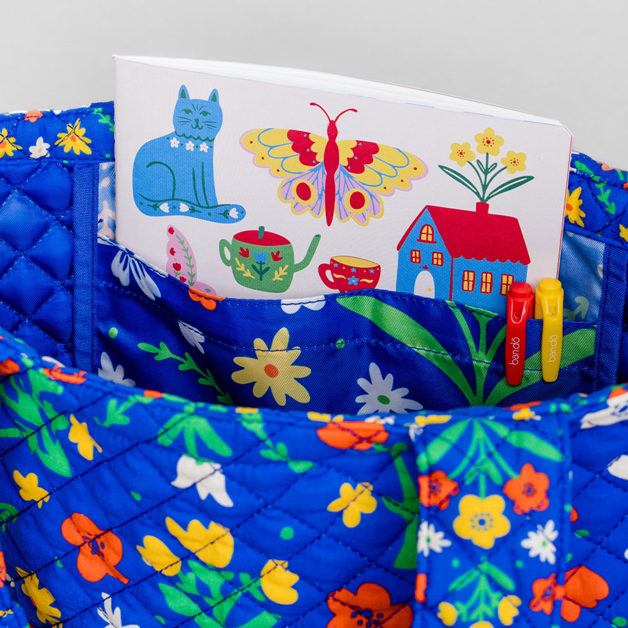 A quilted blue fabric organizer pouch with a colorful floral print, holding a white children's activity book and two small markers.