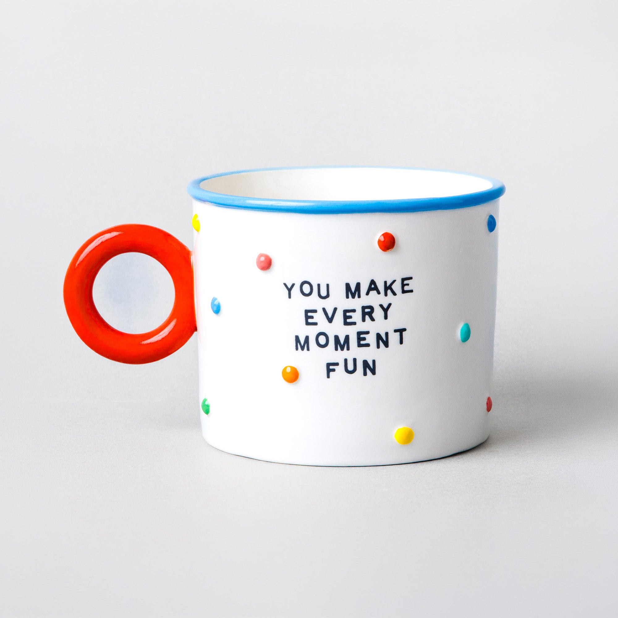A white mug with a bright red handle, blue rim, and multicolored polka dots, featuring the printed text 'YOU MAKE EVERY MOMENT FUN' on the front, set against a light gray background.