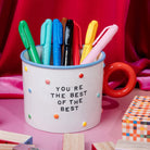 A white ceramic mug with a red handle, decorated with colorful dots and the text 'YOU'RE THE BEST OF THE BEST'. It is filled with various colored pens and is set against a vibrant pink and red background with wooden blocks.