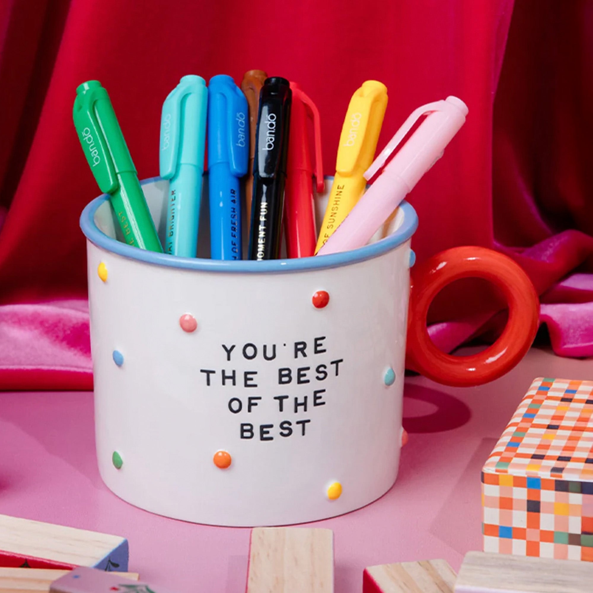 A white ceramic mug with a red handle, decorated with colorful dots and the text 'YOU'RE THE BEST OF THE BEST'. It is filled with various colored pens and is set against a vibrant pink and red background with wooden blocks.