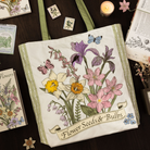 Tote bag with floral design and text on a wooden surface with seed packets and flowers.