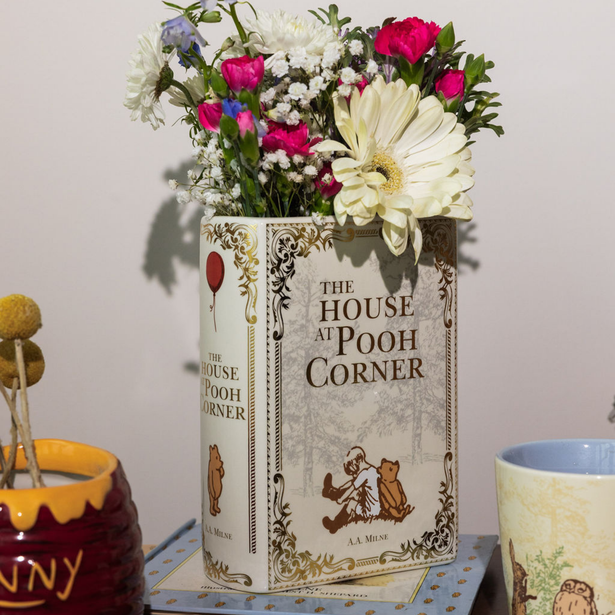 Floral arrangement in a 'The House at Pooh Corner' book on a table with a mug and jar.