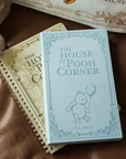 Book titled 'The House at Pooh Corner' by A.A. Milne on a textured surface with a teddy bear and another book.