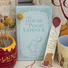 Book titled 'The House at Pooh Corner' with a bookmark featuring a bear and text, surrounded by decorative items.