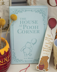 Book titled 'The House at Pooh Corner' with a bookmark featuring a bear and text, surrounded by decorative items.