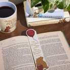 Open book with a bookmark featuring a bear holding a balloon, next to a mug with a bear design on a wooden surface.