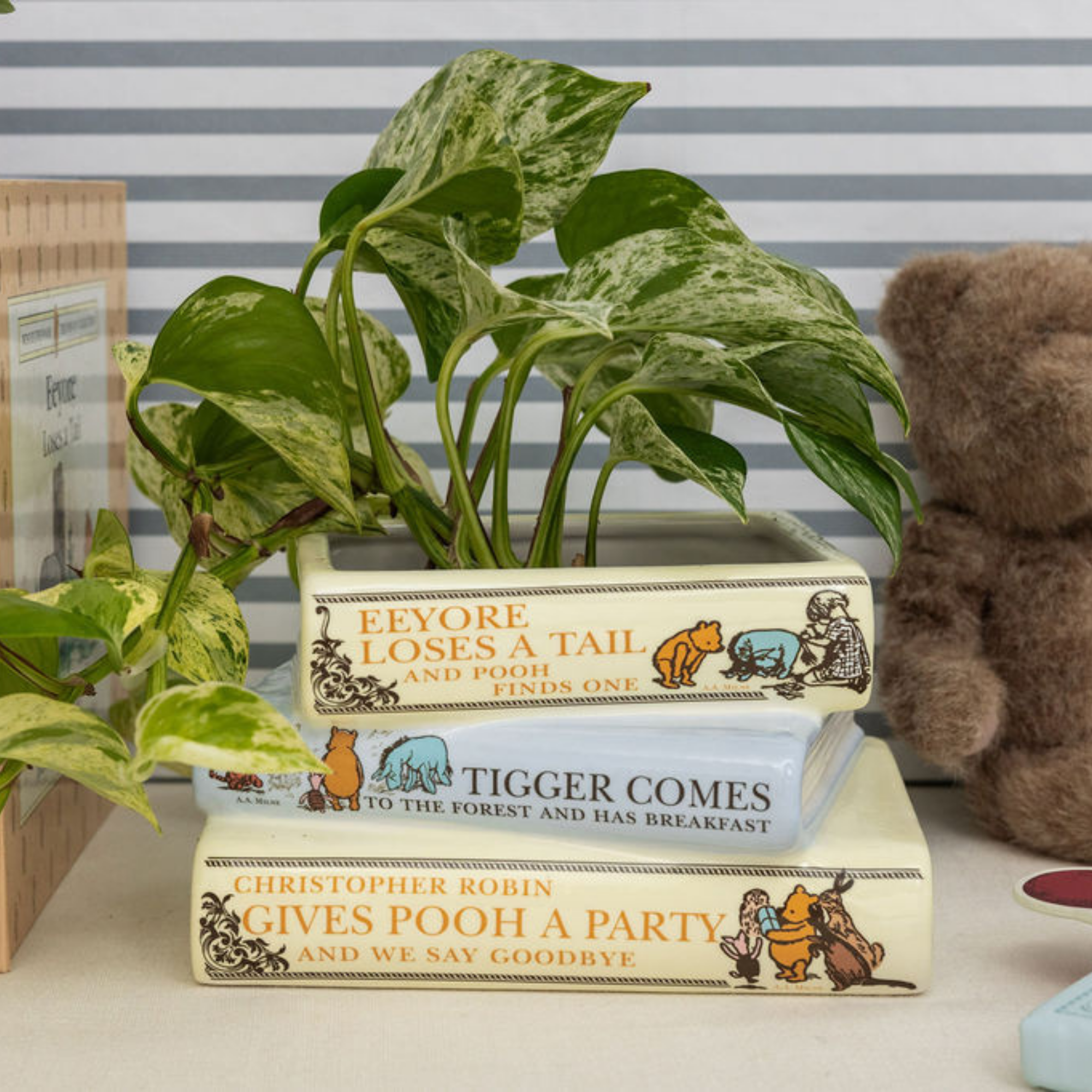 Stack of Winnie the Pooh books with a plant on top against a striped wall.