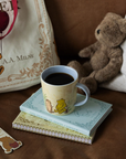 Cup of coffee with a teddy bear and books on a wooden surface, featuring A.A. Milne's 'Winnie the Pooh' books.