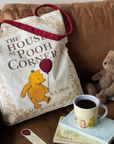 Tote bag with Winnie the Pooh design on a couch with a cup of coffee and teddy bear.