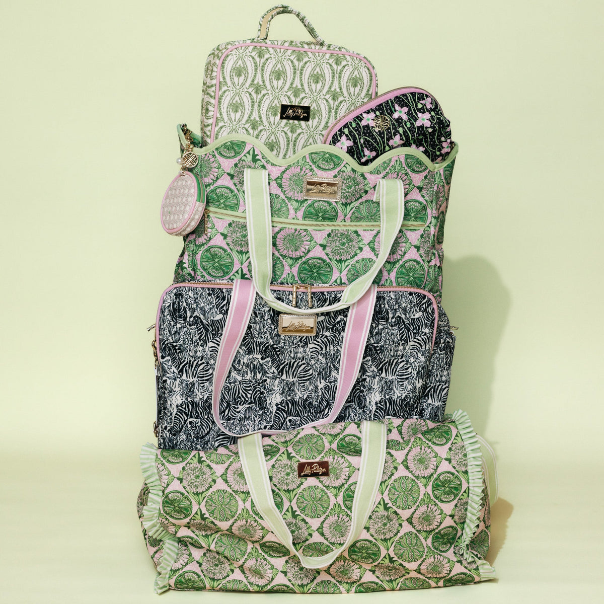 A stack of three patterned fabric bags in green, pink, and black, including a backpack, tote bag, and duffel bag, set against a pale yellow-green background.