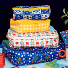 A stack of four colorful zippered packing cubes with various patterns including gingham, floral, and geometric, displayed on a light blue surface against a dark background.