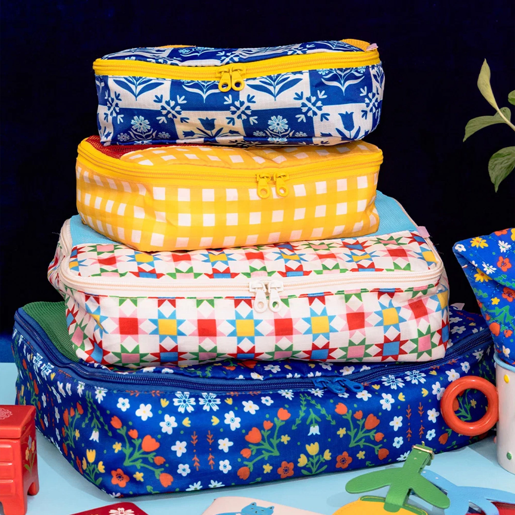 A stack of four colorful zippered packing cubes with various patterns including gingham, floral, and geometric, displayed on a light blue surface against a dark background.