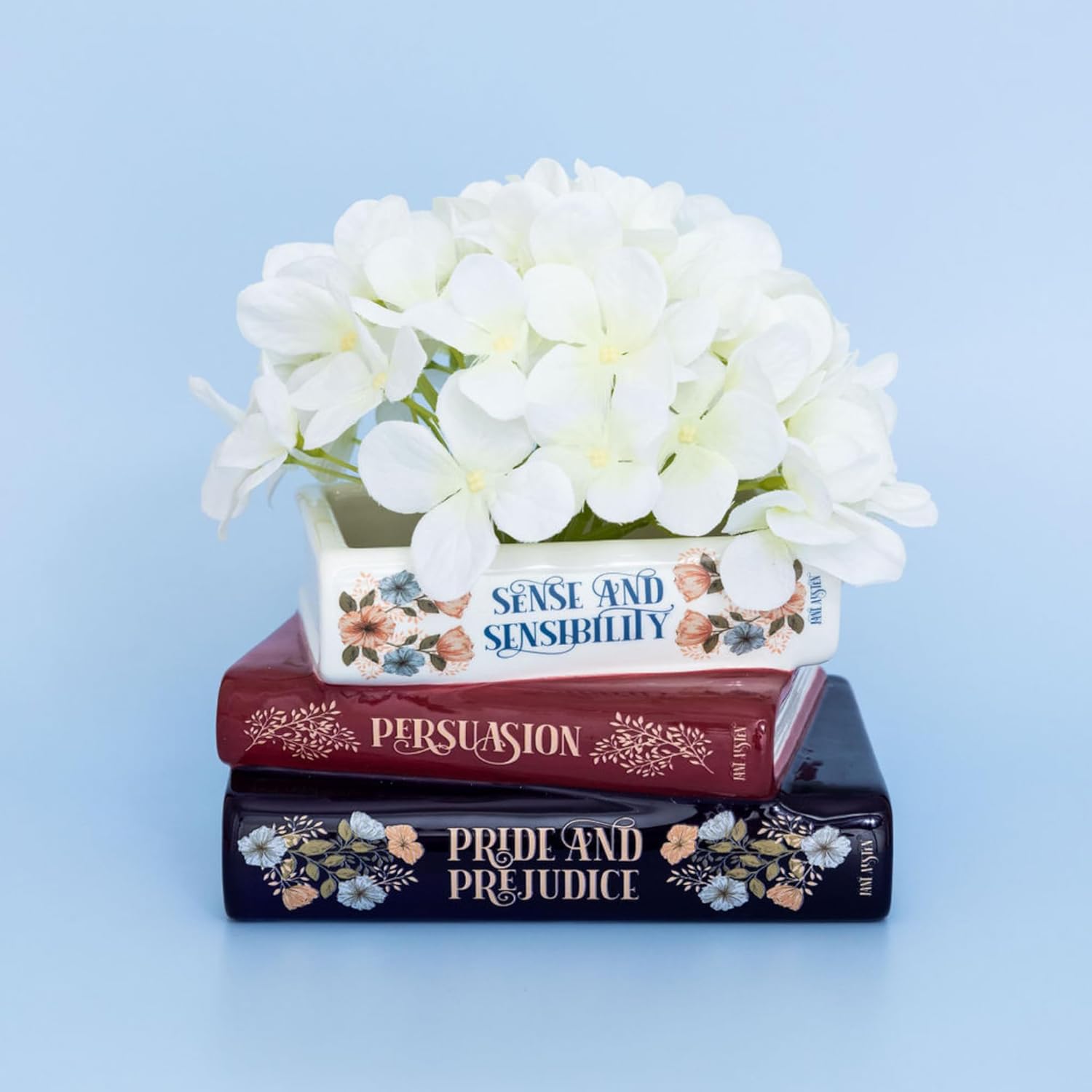 Decorative pot with white flowers on a stack of books titled 'Sense and Sensibility', 'Persuasion', and 'Pride and Prejudice' against a light blue background.