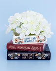 Decorative pot with white flowers on a stack of books titled 'Sense and Sensibility', 'Persuasion', and 'Pride and Prejudice' against a light blue background.