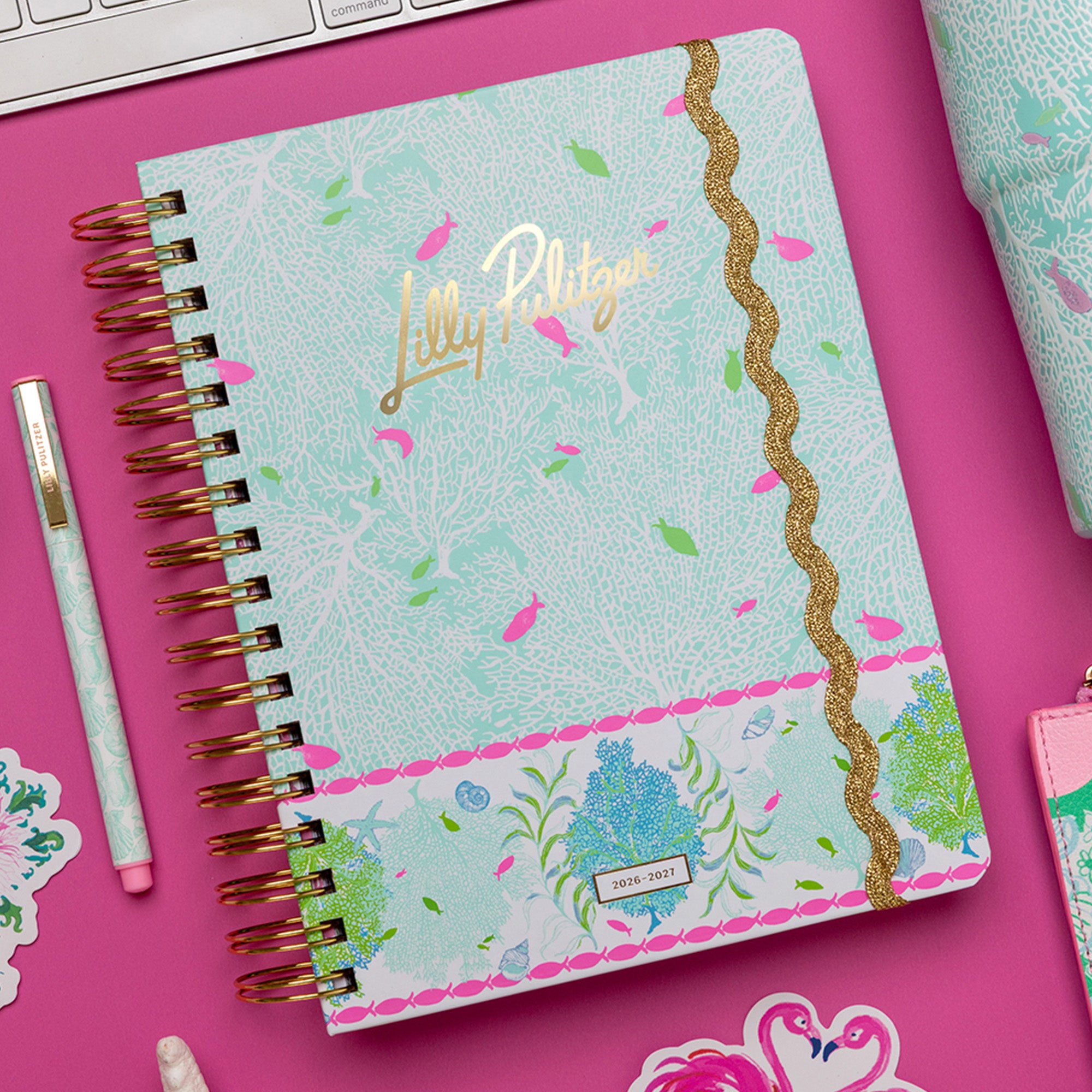 A spiral-bound planner with a light aqua patterned cover, pink and green floral accents, and gold foil script reading 'Lilly Pulitzer.' It is photographed on a bright pink surface with a matching pen, stickers, and part of another item visible around it.
