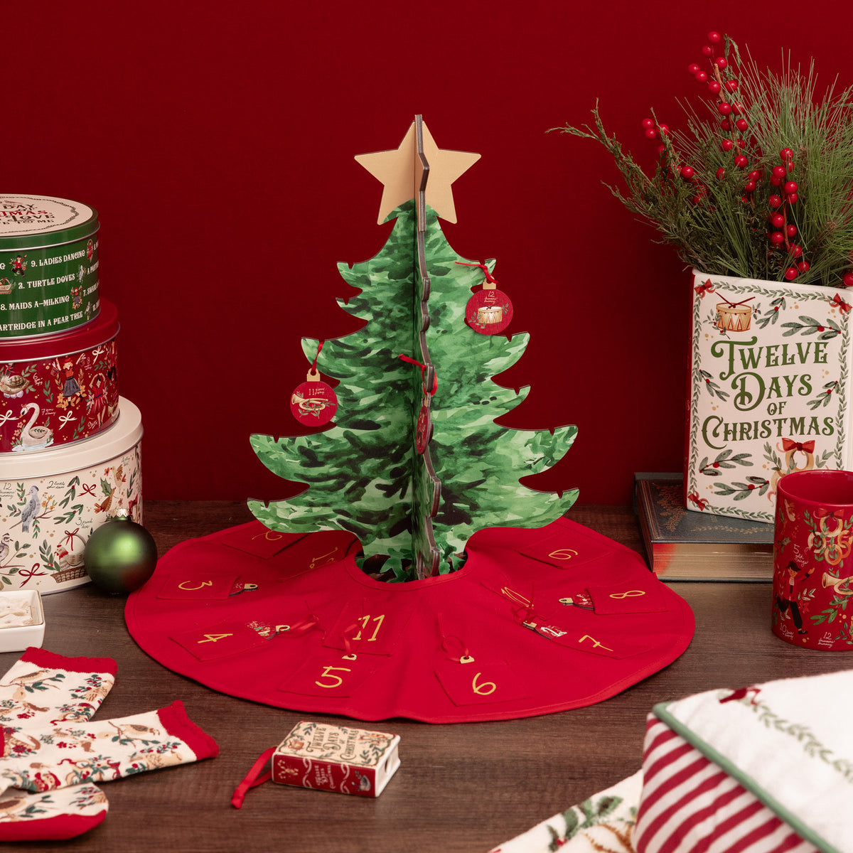 A festive Christmas advent tree set displayed on a dark wooden table against a deep red background. The set includes a green patterned Christmas tree with a gold star topper, a red advent skirt with numbered doors, a small red gift box, and matching stockings.