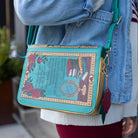 Person wearing a decorative book-shaped handbag with floral and literary design.