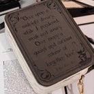 A close-up of a brown faux-leather book bag with gothic-style printed text on the front, resting on an open book with a gold-tone ring pull visible on the right.
