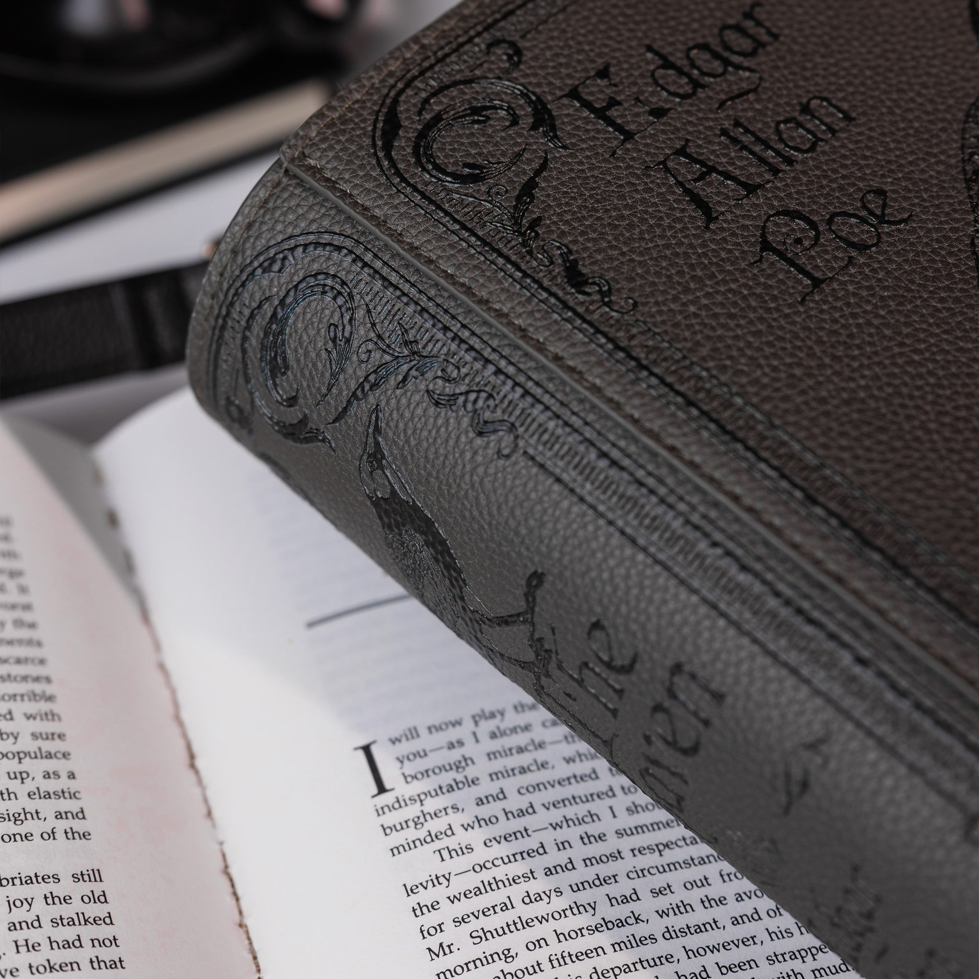 A close-up of a dark brown textured crossbody, showing the embossed title 'The Raven' on the cover, resting on an open book.