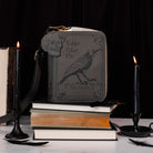 A dark gray faux-leather crossbody bag styled on a stack of books, flanked by lit black candles and a feather, against a black background. The bag features an embossed raven and the text 'Edgar Allan Poe' and 'The Raven'.