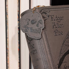 A close-up of a gray faux-leather crossbody featuring a skull-shaped zipper pull, embossed gothic lettering, and a gold zipper pull, set against a background of books.