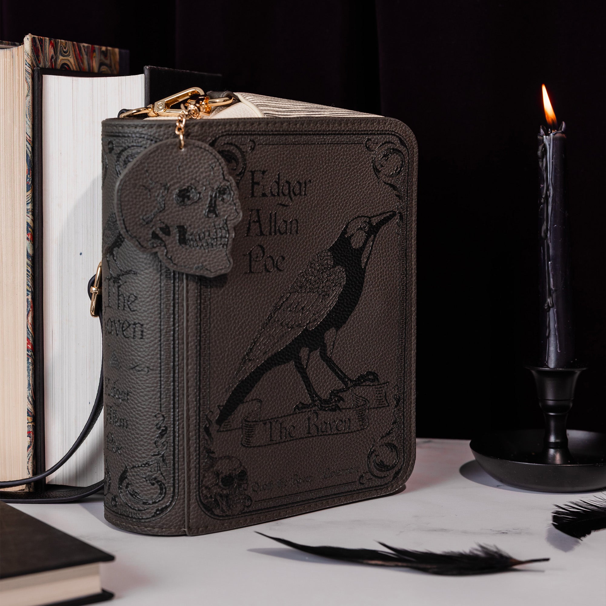 A dark brown faux-leather book-style crossbody with 'The Raven' Edgar Allan Poe design, styled on a tabletop with books, a lit candle, and feathers against a black background.
