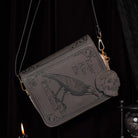 A dark gray faux-leather crossbody bag styled like an old book cover, featuring embossed gothic text, a raven illustration, and gold-tone hardware, shown against a dark draped background.