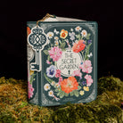 A decorative book-shaped handbag styled like 'The Secret Garden' book cover, featuring a floral wreath illustration and a large ornate key charm, displayed on moss against a black background.