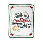 A square white embroidered pillow or cushion with a green border and festive text reading 'On the first day of Christmas, my true love gave to me,' decorated with red bows, green leaves, and small holiday illustrations.