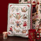 A rectangular decorative pillow with a Christmas-themed illustrated calendar design, styled on a dark wooden surface next to a lit candle, a red mug, and a matching patterned blanket against a dark red background.