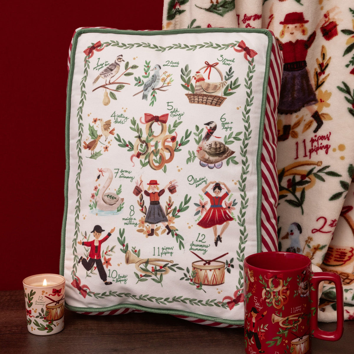 A rectangular decorative pillow with a Christmas-themed illustrated calendar design, styled on a dark wooden surface next to a lit candle, a red mug, and a matching patterned blanket against a dark red background.