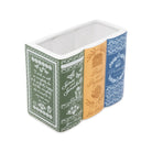 A rectangular ceramic container with a three-spine design featuring vintage book covers. The left spine is dark green with white script and botanical illustrations, the middle spine is warm orange with gold-toned text and a house illustration, and the right spine is blue with white floral patterns.