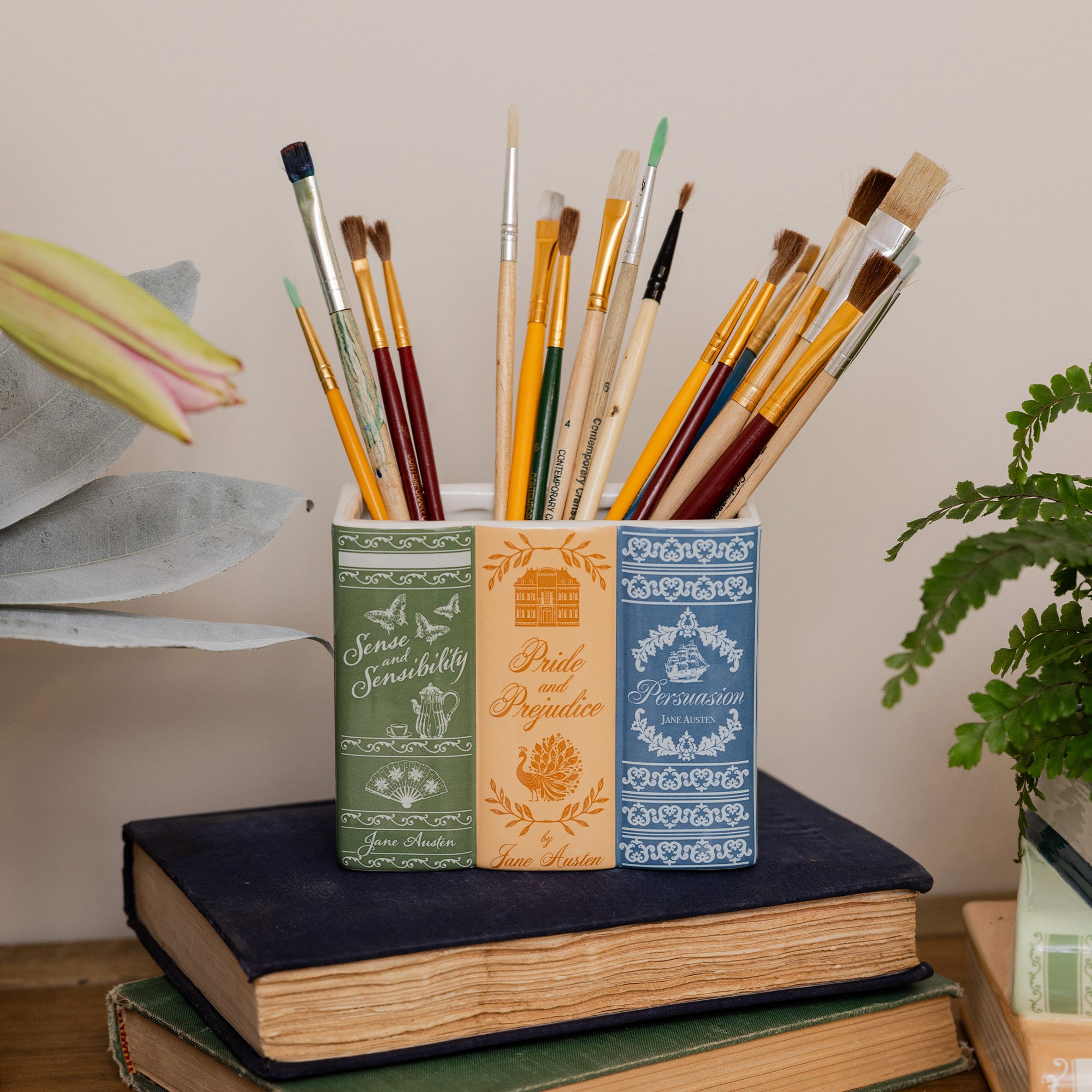 A decorative holder shaped like three vintage book spines in green, orange, and blue, filled with artist paintbrushes and styled on top of old books with plants in the background.