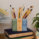 A decorative holder shaped like three vintage book spines in green, orange, and blue, filled with artist paintbrushes and styled on top of old books with plants in the background.
