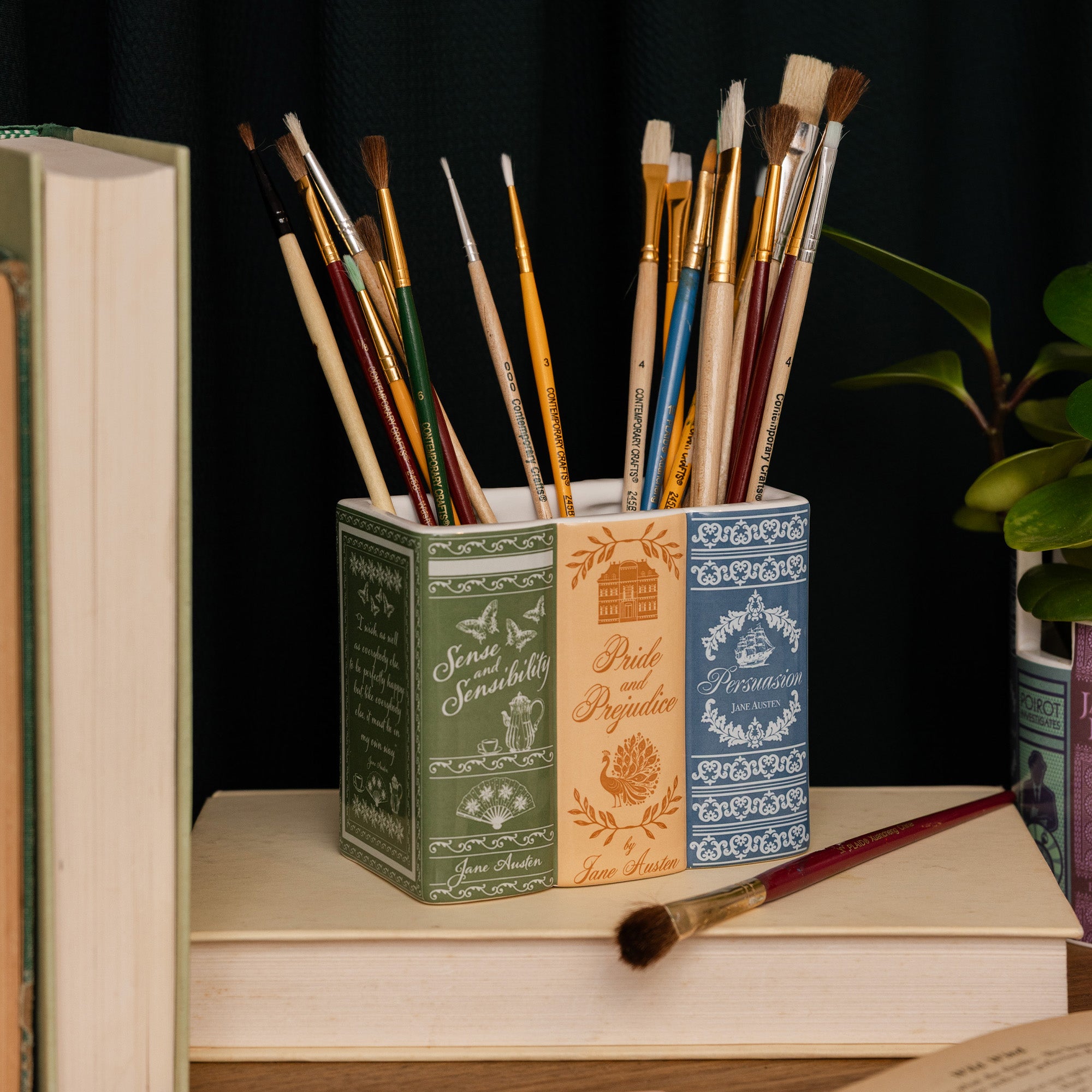 A rectangular ceramic container with a three-spine design featuring vintage book covers. The left spine is dark green with white script and botanical illustrations, the middle spine is warm orange with gold-toned text and a house illustration, and the right spine is blue with white floral patterns.