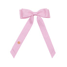 A light pink satin bow with long tails, featuring a small round gold-toned decorative emblem on one of the tails, shown against a plain white background.