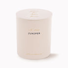 A cylindrical candle in a matte cream-colored jar with a matching lid, featuring gold and black text on the front and lid. The brand 'Amélin Archive' is printed at the bottom, and the scent 'Juniper' is labeled in black.