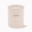 A matte blush pink glass candle jar with a white wax fill and a single wick. The front features black text reading 'JUNIPER' with pink and gold foil accents above and below, on a clean white background.