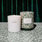 A white glass candle jar labeled 'Amelin Archive Juniper' with a small flame, standing beside its patterned green and white cylindrical box on a matching patterned surface in front of a dark green velvet curtain.
