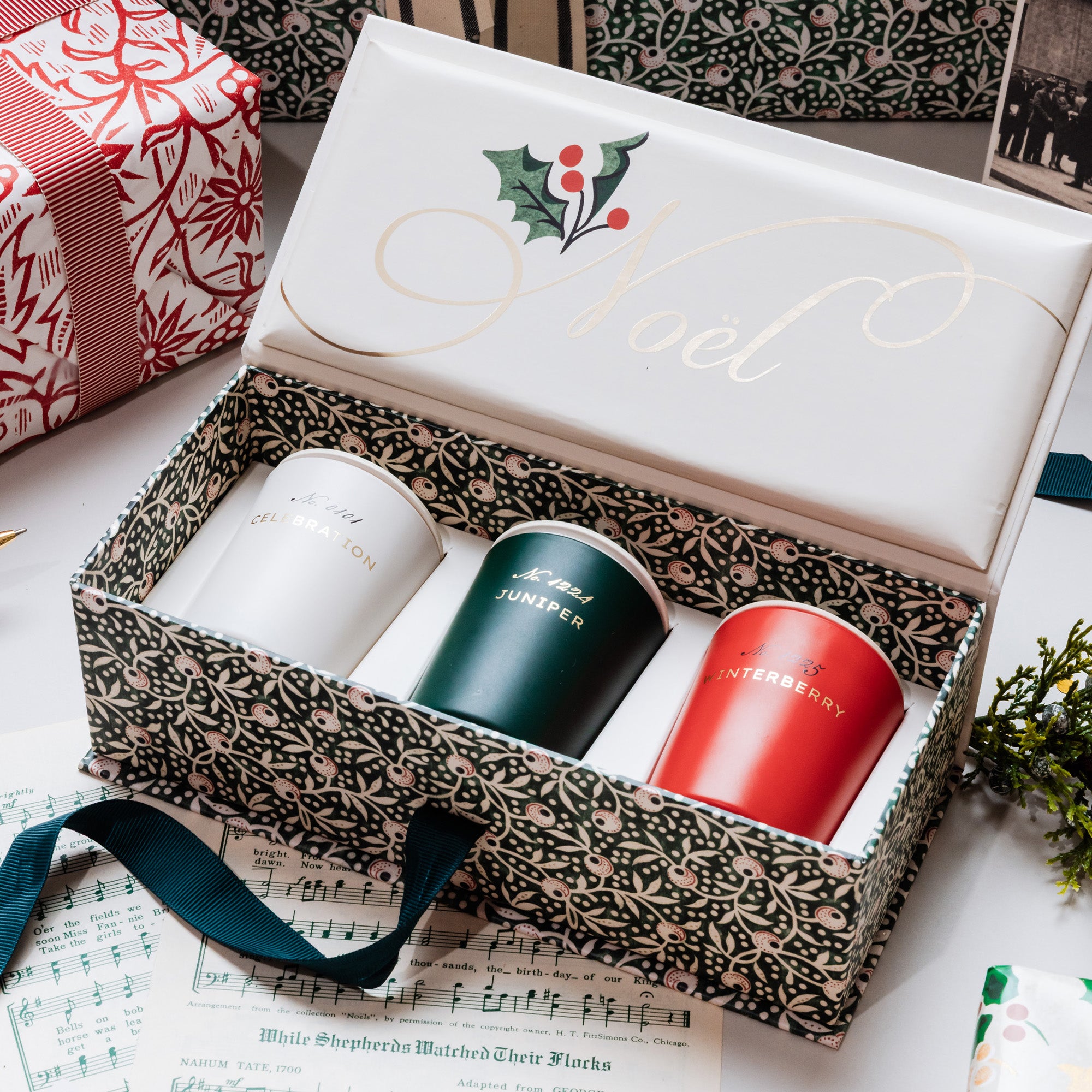 A boxed set of three Christmas-themed candles displayed in an open decorative gift box. The box lid is white with gold cursive script reading 'Noel' and a holly graphic.