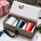 A boxed set of three Christmas-themed candles displayed in an open decorative gift box. The box lid is white with gold cursive script reading 'Noel' and a holly graphic.