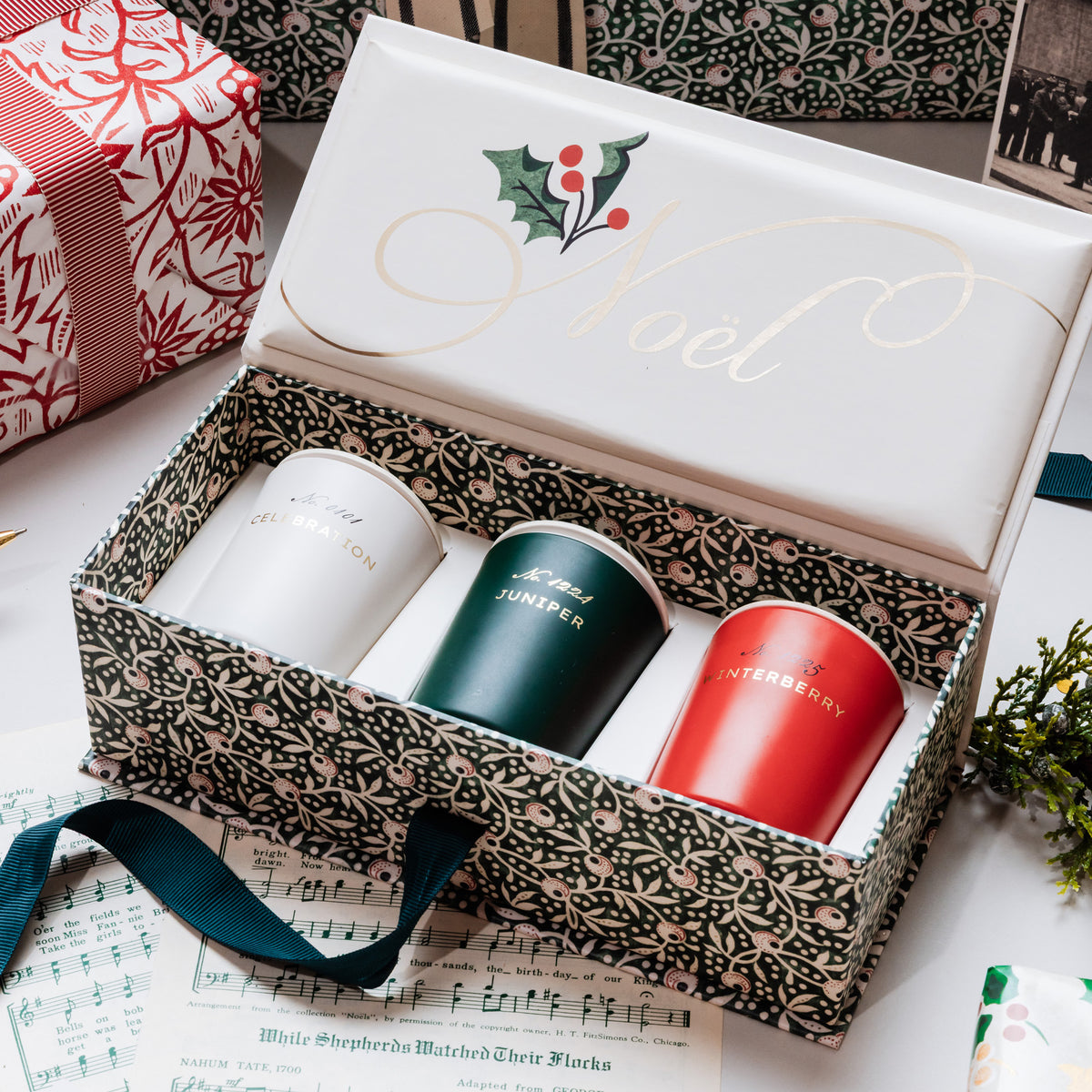 A boxed set of three Christmas-themed candles displayed in an open decorative gift box. The box lid is white with gold cursive script reading 'Noel' and a holly graphic.
