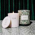 lit Amëlin Archive Juniper scented candle in a cylindrical glass container with a green and white botanical pattern packaging and a white label in the center.