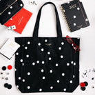 A black canvas tote bag with white polka dots by Kate Middleton, styled in a flat-lay arrangement with a black 2027 spiral notebook, a red notebook, eyeglasses, and other desk accessories on a white background.