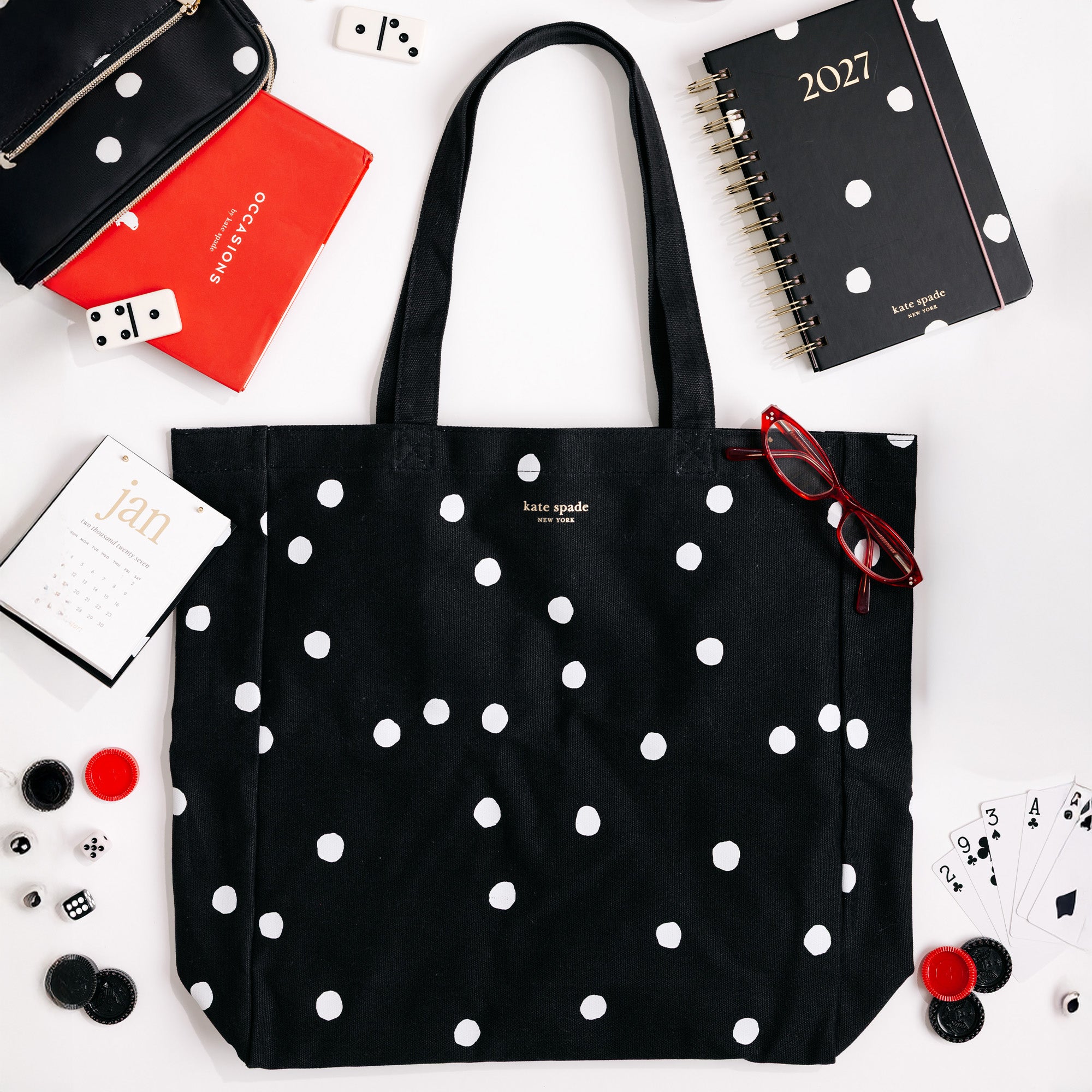 A black canvas tote bag with white polka dots by Kate Middleton, styled in a flat-lay arrangement with a black 2027 spiral notebook, a red notebook, eyeglasses, and other desk accessories on a white background.
