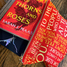 A red zippered fabric pouch printed with white and yellow text from the book 'Thorns and Roses' by Sarah J. Maas, shown open on a wooden surface next to the book.