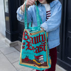 Person holding a 'Beauty and the Beast' book-themed bag on a sidewalk.